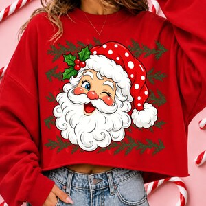 May include: A red long-sleeve sweatshirt featuring a graphic of a winking Santa Claus wearing a red hat with white polka dots. The design includes green holly leaves with red berries and a repeating pattern of green pine branches in the background.