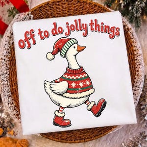May include: A white t-shirt featuring a graphic of a goose wearing a red and green patterned sweater, a matching Santa hat, and red boots. The text above the goose reads "off to do jolly things" in a red, distressed, rounded font style.