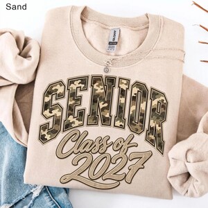 May include: A sand-coloured crewneck sweatshirt featuring the word SENIOR in large, block-style letters with a green, brown, and tan camouflage pattern. Below, the text Class of 2027 is printed in a cursive font with a dark brown outline.
