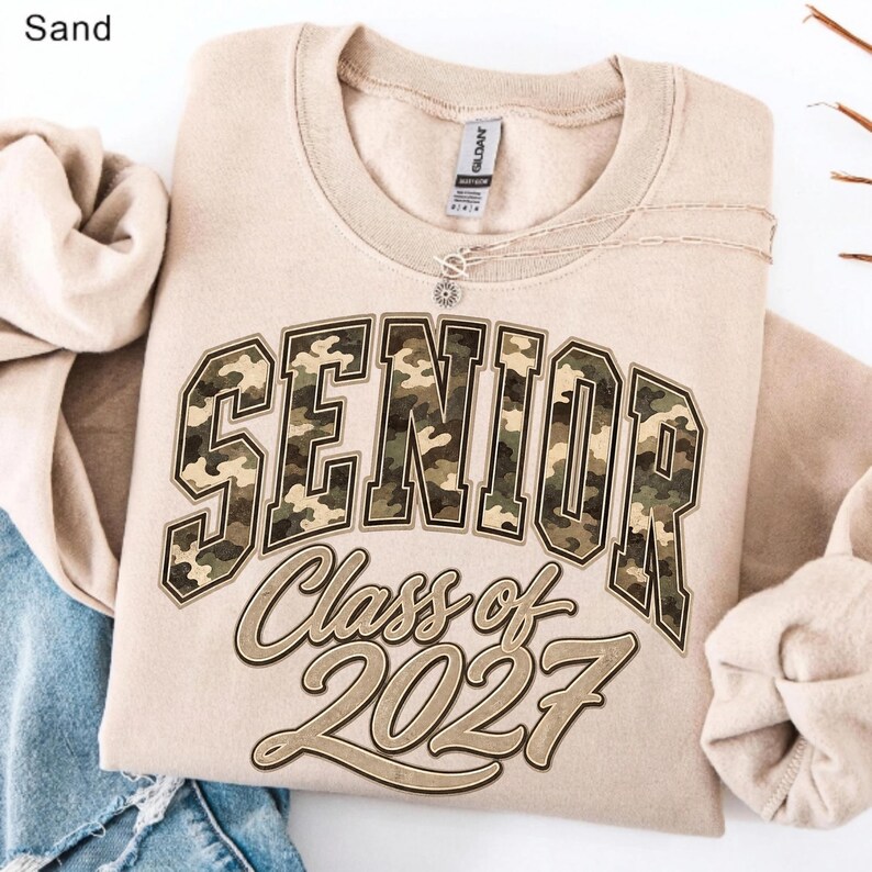 May include: A sand-coloured crewneck sweatshirt featuring the word SENIOR in large, block-style letters with a green, brown, and tan camouflage pattern. Below, the text Class of 2027 is printed in a cursive font with a dark brown outline.
