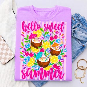 May include: A light purple t-shirt featuring a graphic print with the text "hello sweet summer" in bright pink script. The design includes illustrations of halved coconuts, yellow lemon slices, red cherries, blue ice cubes, and colorful cocktail umbrellas.