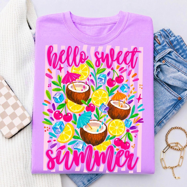 May include: A light purple t-shirt featuring a graphic print with the text "hello sweet summer" in bright pink script. The design includes illustrations of halved coconuts, yellow lemon slices, red cherries, blue ice cubes, and colorful cocktail umbrellas.
