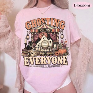 Pode incluir: Uma t-shirt rosa claro com uma impress&atilde;o gr&aacute;fica de um fantasma usando um turbante atr&aacute;s de uma bola de cristal. O texto diz GHOSTING EVERYONE, SPIRIT LINE e NO APPOINTMENTS NO APPARITIONS. O design inclui um gato preto, livros e uma ab&oacute;bora.