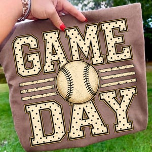 May include: A brown t-shirt featuring the text "GAME DAY" in large, cream-coloured block letters with a black polka dot pattern. A central graphic of a baseball replaces the letter "A" in "GAME" and the letter "D" in "DAY" is stylised.
