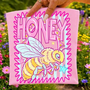 May include: A light pink fabric item featuring the word HONEY in large, bold, dark pink letters. Below the text is a yellow and black striped bee with light blue wings and legs, surrounded by small yellow and blue stars on a striped background.