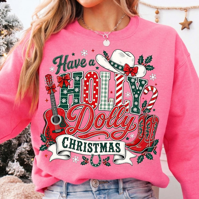 May include: A bright pink sweatshirt featuring a graphic print with the text Have a HOLLY Dolly CHRISTMAS. The design includes a white cowboy hat, a red acoustic guitar, red cowboy boots, a candy cane, holly leaves, and snowflakes.