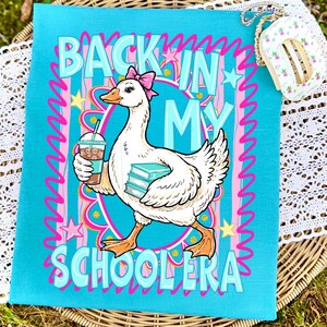 May include: A teal-colored graphic tee featuring a cartoon goose wearing a pink bow and carrying books and a coffee drink. The text reads "BACK IN MY SCHOOLERA" in a playful font, surrounded by stars and swirls.