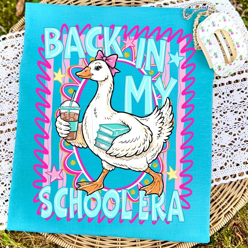 May include: A teal-colored graphic tee featuring a cartoon goose wearing a pink bow and carrying books and a coffee drink. The text reads "BACK IN MY SCHOOLERA" in a playful font, surrounded by stars and swirls.