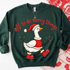 May include: A dark green crewneck sweatshirt featuring a graphic of a white goose wearing a red Santa hat, a red vest with white trim, and red boots. The text "off to do merry things" is printed in red above the goose, with small holly sprigs.
