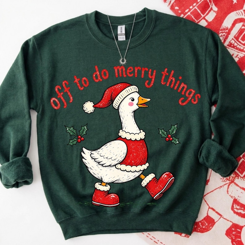 May include: A dark green crewneck sweatshirt featuring a graphic of a white goose wearing a red Santa hat, a red vest with white trim, and red boots. The text "off to do merry things" is printed in red above the goose, with small holly sprigs.