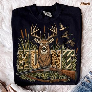 May include: A black t-shirt featuring a graphic of a deer head centred above the word HUNT in block letters. The letters are filled with camouflage and leopard patterns. A shotgun is positioned below the text, with three flying ducks above.