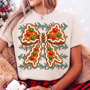 Op de afbeelding: Een cr&egrave;mekleurig t-shirt met een grote afbeelding van een peperkoekstrik versierd met wit glazuur en rode en groene snoepjes. De strik is omringd door een patroon van groene dennentakken en kleine rode kerstlichtjes.
