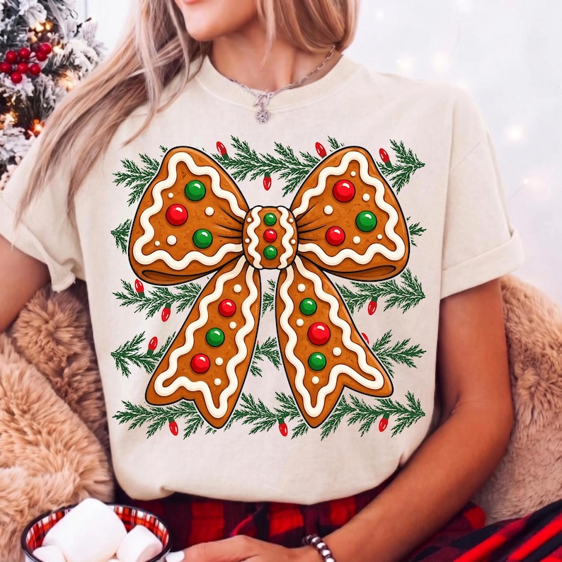 Op de afbeelding: Een cr&egrave;mekleurig t-shirt met een grote afbeelding van een peperkoekstrik versierd met wit glazuur en rode en groene snoepjes. De strik is omringd door een patroon van groene dennentakken en kleine rode kerstlichtjes.