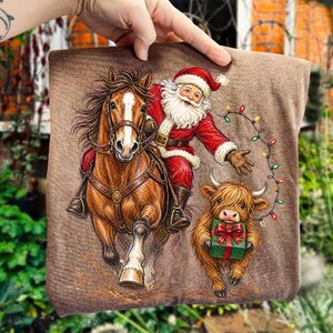 Puede incluir: Una camiseta marr&oacute;n con una ilustraci&oacute;n gr&aacute;fica de Santa Claus montando un caballo marr&oacute;n con una mancha blanca en la cara. Junto a ellos, una peque&ntilde;a y peluda vaca Highland marr&oacute;n lleva un regalo verde con un lazo rojo, rodeado de luces.