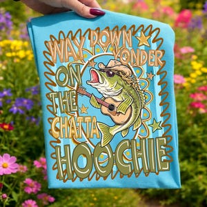 May include: A light blue t-shirt featuring a cartoon bass fish wearing a cowboy hat and sunglasses, playing a guitar. The shirt has the text "Way Down Yonder On The Chatta Hoochie" in a decorative font.