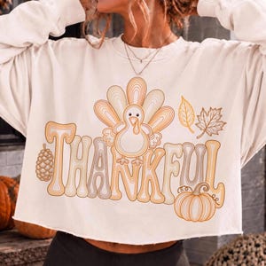 May include: A cream-colored long-sleeve sweatshirt featuring a graphic print with the word THANKFUL in large, outlined letters. The design includes a central turkey, a pinecone, two autumn leaves, and a pumpkin, all rendered in orange and brown.
