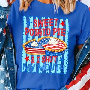 Op de afbeelding: Een koningsblauw t-shirt met een afbeelding van twee taarten met Amerikaanse vlagpatronen en slagroom. De tekst SWEET POTATO PIE & I SHUT MY MOUTH staat in rode en blauwe letters, omringd door rode en blauwe sterren op een gestreepte achtergrond.