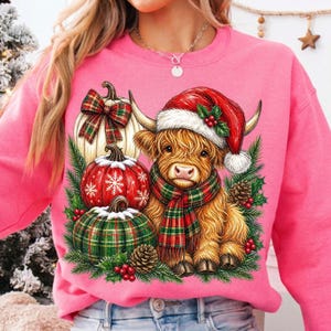 May include: A bright pink sweatshirt featuring a graphic of a fluffy Highland cow wearing a red Santa hat and a plaid scarf, surrounded by three pumpkins with plaid and snowflake patterns, pine branches, pinecones, and red holly berries.