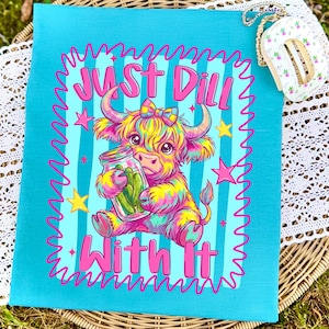 Puede incluir: Una camiseta de color turquesa brillante con una colorida ilustraci&oacute;n de una vaca Highland estilo tie-dye sosteniendo un frasco de pepinillos. El gr&aacute;fico incluye el texto "Just Dill With It" en rosa, rodeado por un borde rosa ondulado y estrellas amarillas.