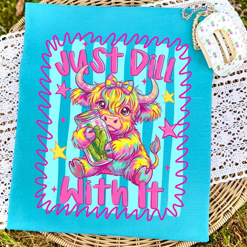 Puede incluir: Una camiseta de color turquesa brillante con una colorida ilustraci&oacute;n de una vaca Highland estilo tie-dye sosteniendo un frasco de pepinillos. El gr&aacute;fico incluye el texto "Just Dill With It" en rosa, rodeado por un borde rosa ondulado y estrellas amarillas.