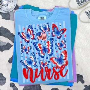 May include: A light blue t-shirt featuring a graphic print of multiple butterflies with red, white, and blue patterns, a small American flag, and the word Nurse in red script. The shirt has a Comfort Colors label at the inner neckline.
