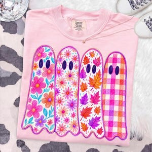 May include: A light pink t-shirt featuring a graphic of four ghosts standing side by side. Each ghost has a unique pattern: the first has colorful flowers, the second has small daisies, the third has autumn leaves, and the fourth has a pink and orange plaid pattern.