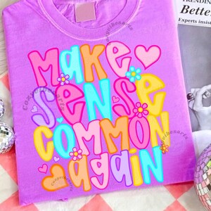 May include: A lilac-colored t-shirt with the phrase "MAKE SENSE COMMON AGAIN" in colorful, bubble-style letters. The design includes hearts and flower accents. The shirt is made of a soft material.