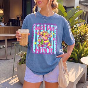 Puede incluir: Una camiseta azul de manga corta con un gr&aacute;fico de una vaca highland colorida sosteniendo un frasco de pepinillos. El dise&ntilde;o incluye un fondo de rayas azul claro, estrellas rosas y el texto Just Dill With It en fuente rosa al frente.