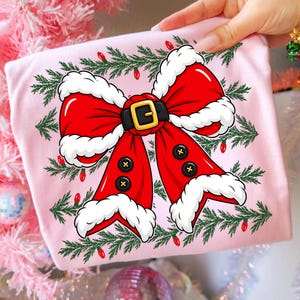 Puede incluir: Una camiseta rosa claro con un gr&aacute;fico de un gran lazo rojo con borde de piel blanca, un cintur&oacute;n negro con hebilla dorada y botones negros. El dise&ntilde;o est&aacute; rodeado por ramas de pino verdes y peque&ntilde;as luces navide&ntilde;as rojas.