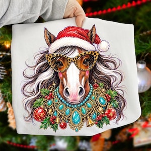 May include: A white t-shirt featuring a graphic illustration of a brown horse wearing a red Santa hat, leopard print sunglasses, and a decorative necklace made of turquoise stones, gold accents, red Christmas ornaments, and green holly leaves.