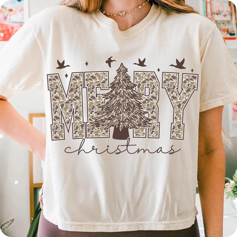 May include: Cream-colored t-shirt with the words "MERRY" in camouflage print and a brown Christmas tree graphic. The word "christmas" is written in a cursive font below the main text. Above the word "MERRY" are three birds.
