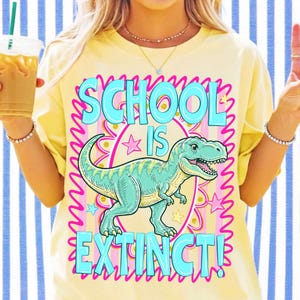 Op de afbeelding: Een lichtgeel t-shirt met een grafische print van een groene dinosaurus in het midden. De tekst luidt SCHOOL IS EXTINCT in grote lichtblauwe letters. Het ontwerp bevat een roze geschulpte rand, kleine roze en gele sterren en gouden stippen.