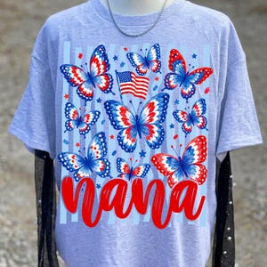 May include: A light gray t-shirt featuring a graphic print of multiple butterflies with red, white, and blue patterns. A small American flag is positioned among the butterflies. The word Nana is printed in large, bold red script at the bottom.
