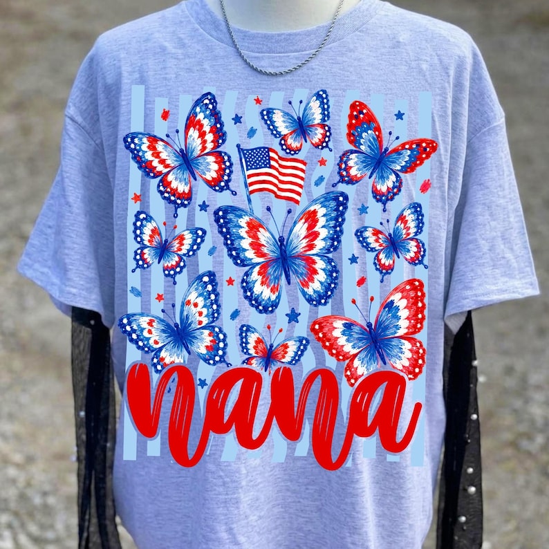 May include: A light gray t-shirt featuring a graphic print of multiple butterflies with red, white, and blue patterns. A small American flag is positioned among the butterflies. The word Nana is printed in large, bold red script at the bottom.