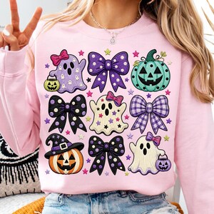 K&ouml;nnte beinhalten: Ein hellrosa Sweatshirt mit Rundhalsausschnitt und einem Gittermuster aus Halloween-Grafiken. Das Design zeigt drei Geister, zwei K&uuml;rbisse und vier Schleifen in Lila, Schwarz und T&uuml;rkis, die mit kleinen bunten Sternen verziert sind.