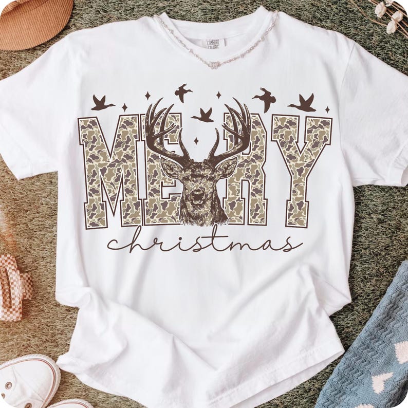 May include: White t-shirt with a "Merry Christmas" graphic. The word "MERRY" is in a camouflage pattern, with a deer head in the center. Above the word "MERRY" are three birds. The word "christmas" is written in a cursive font.