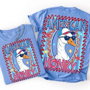 May include: A light blue t-shirt featuring a graphic of a white goose wearing a blue and white baseball cap and black sunglasses. The text "AMERICAN HONRY" is printed in blue and red, surrounded by a red wavy border and yellow stars.