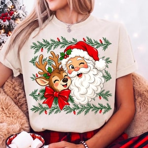 May include: A cream-colored t-shirt featuring a graphic of Santa Claus and a reindeer with a large red bow. The design is framed by green pine branches with small red lights. Santa wears a red hat and the reindeer has brown antlers.