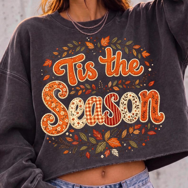 May include: A charcoal gray cropped sweatshirt featuring the text 'Tis the Season' in large, orange, patterned letters. The design is surrounded by a circular arrangement of colorful autumn leaves and small floral accents in shades of orange and green.
