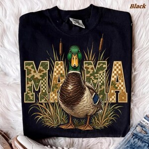 May include: A black t-shirt featuring a graphic of a mallard duck standing in tall grass. The word MAMA is printed in large, block letters with a green and brown camouflage pattern. Two cattails are positioned vertically behind the duck.