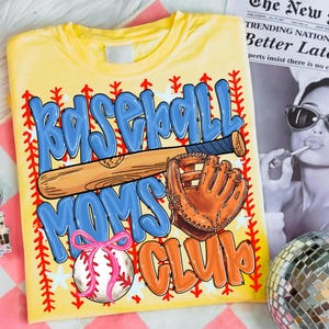 May include: Yellow t-shirt with a baseball-themed graphic. The design features the words "Baseball Moms Club" in blue and orange bubble letters, along with a baseball bat, glove, and a baseball with a pink bow. Red stitching and white stars border the design.
