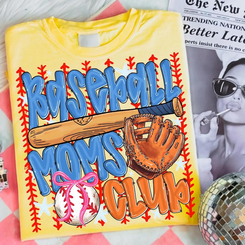 May include: Yellow t-shirt with a baseball-themed graphic. The design features the words "Baseball Moms Club" in blue and orange bubble letters, along with a baseball bat, glove, and a baseball with a pink bow. Red stitching and white stars border the design.