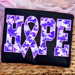 May include: A black t-shirt featuring the word HOPE in large block letters. The letter O is replaced by an awareness ribbon. The letters and ribbon have a purple, light purple, and white camouflage pattern with a subtle speckled texture.