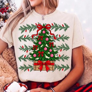 Może przedstawiać: Kremowy t-shirt z grafiką zielonej choinki w drewnianej skrzynce. Drzewko jest ozdobione r&oacute;żowymi bombkami i czerwonymi kokardami, a na szczycie znajduje się duża czerwona kokarda. Zielone gałązki sosny i czerwone żar&oacute;wki otaczają choinkę.