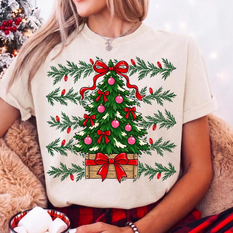 Może przedstawiać: Kremowy t-shirt z grafiką zielonej choinki w drewnianej skrzynce. Drzewko jest ozdobione r&oacute;żowymi bombkami i czerwonymi kokardami, a na szczycie znajduje się duża czerwona kokarda. Zielone gałązki sosny i czerwone żar&oacute;wki otaczają choinkę.