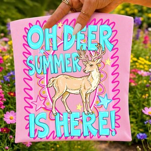 Może przedstawiać: Jasnor&oacute;żowy przedmiot z tkaniny z centralną ilustracją beżowego jelenia stojącego w ozdobnej owalnej ramce. Napis "OH DEER SUMMER IS HERE!" jest wydrukowany dużymi, jasnoniebieskimi literami. Wz&oacute;r zawiera r&oacute;żowe faliste obw&oacute;dki i niebieskie gwiazdki.