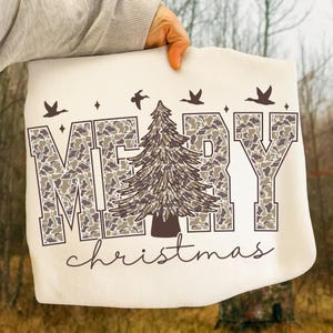 May include: Cream-colored tote bag with the words "MERRY christmas" in a camouflage pattern. A brown Christmas tree is in the center, with brown birds and stars above the text. The bag is held up by a person.