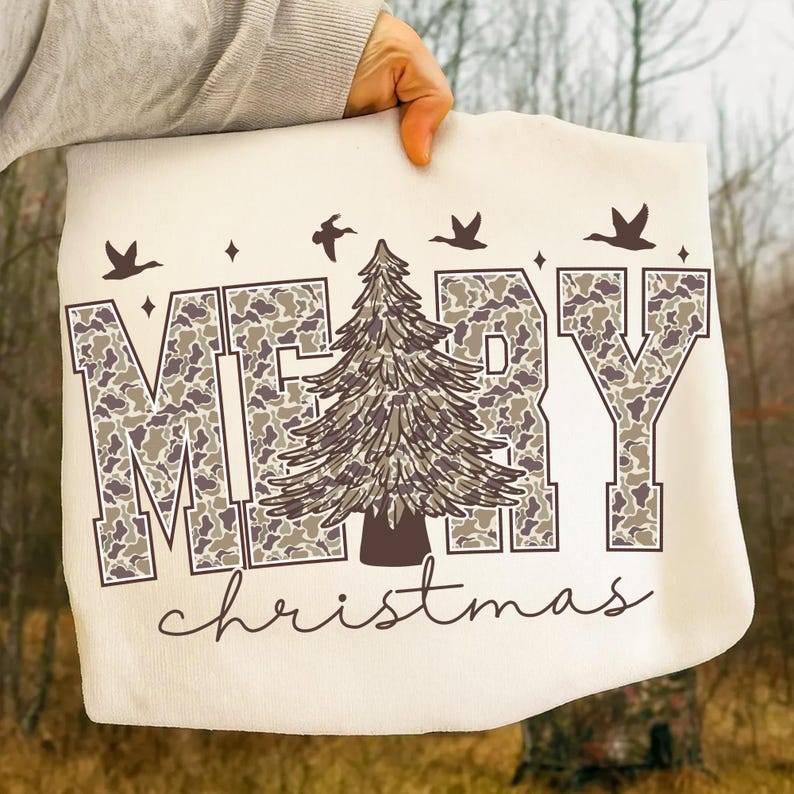 May include: Cream-colored tote bag with the words "MERRY christmas" in a camouflage pattern. A brown Christmas tree is in the center, with brown birds and stars above the text. The bag is held up by a person.