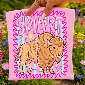 May include: A light pink fabric item featuring a graphic illustration of a brown bison standing in the centre. Above the bison, the word SMART is printed in large pink letters. The design includes a bright blue striped background and pink stars.