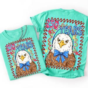 Puede incluir: Una camiseta verde menta con un gr&aacute;fico de un &aacute;guila calva con una pajarita azul. El texto 250 YEARS est&aacute; impreso sobre el &aacute;guila en azul y rojo. El dise&ntilde;o est&aacute; enmarcado por un borde rojo ondulado con peque&ntilde;as estrellas azules y amarillas.